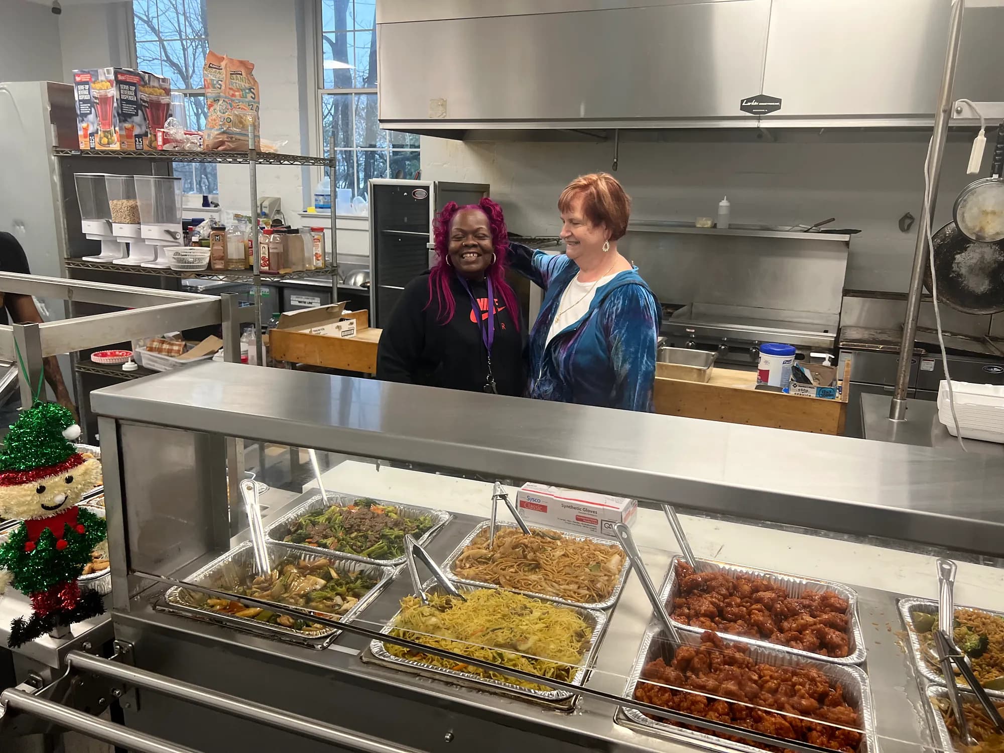Two smiling women stand behind a steam table of General Tso's noodles, beef and broccoli and orange chicken in the Covenant House kitchen