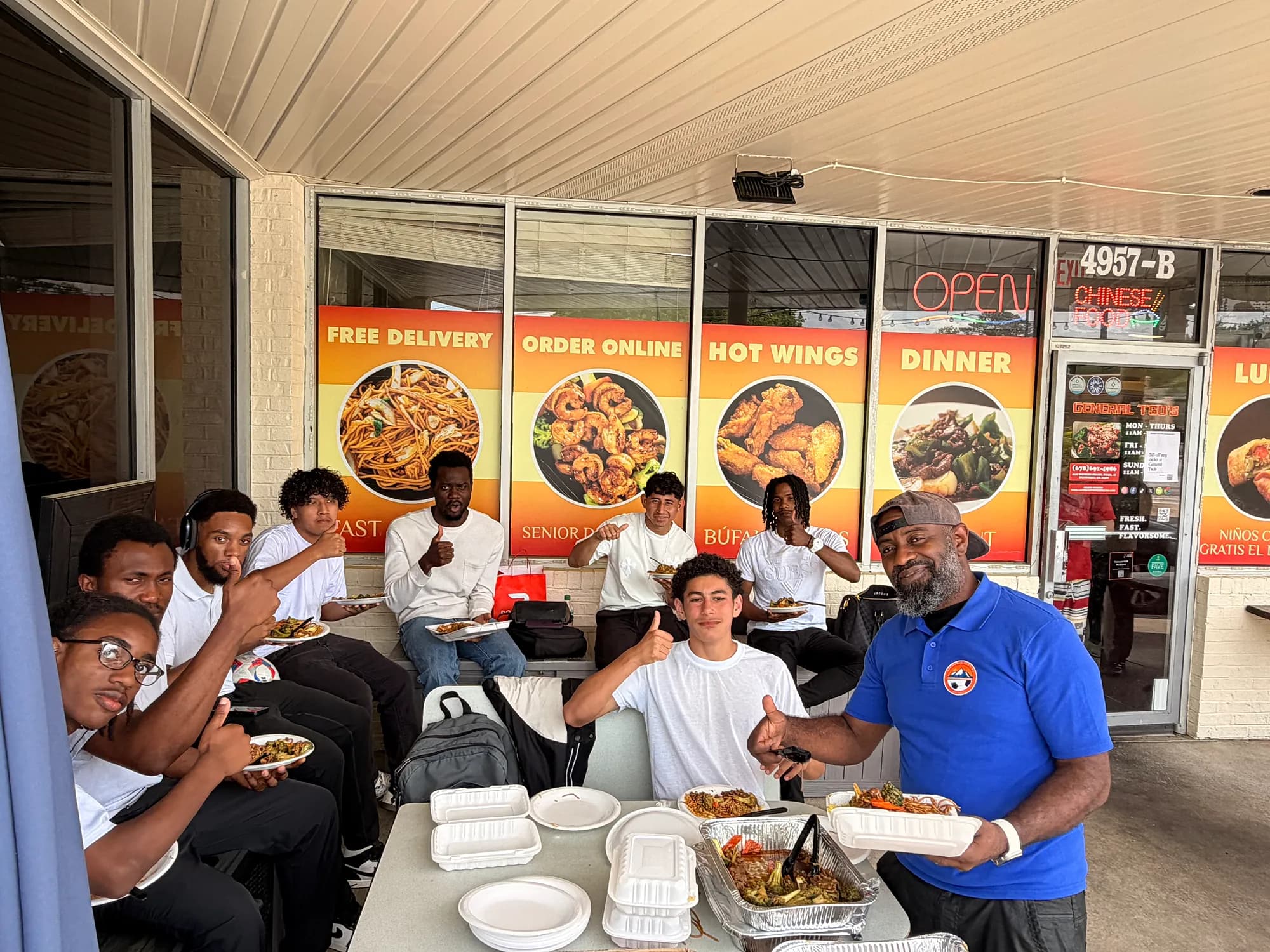 A man in a blue polo serves trays of food to Lithonia City FC players, who give a thumbs-up on the General Tso's patio