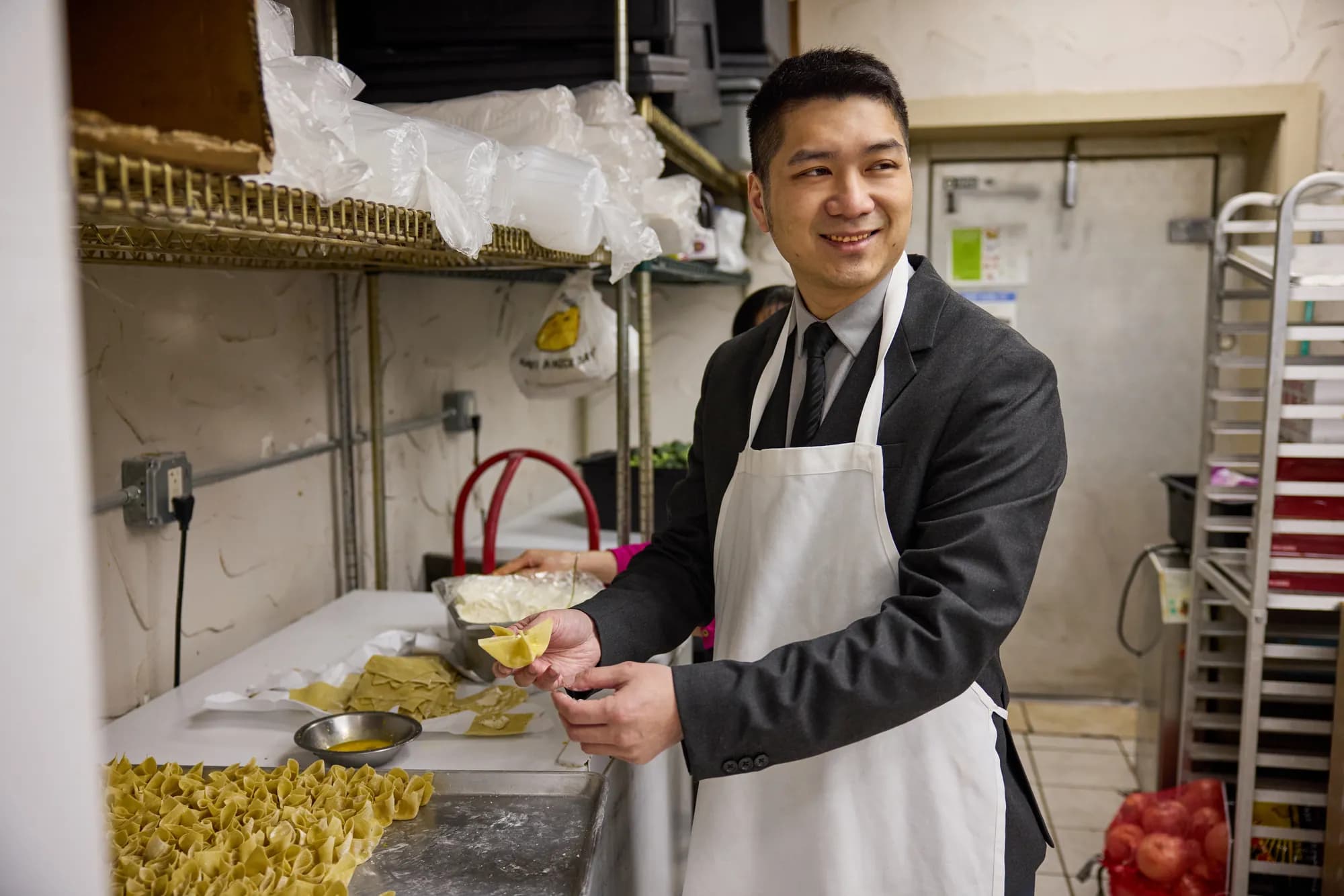 Patrick Cheng, in an apron over his suit, smiles while folding wontons at a kitchen prep table