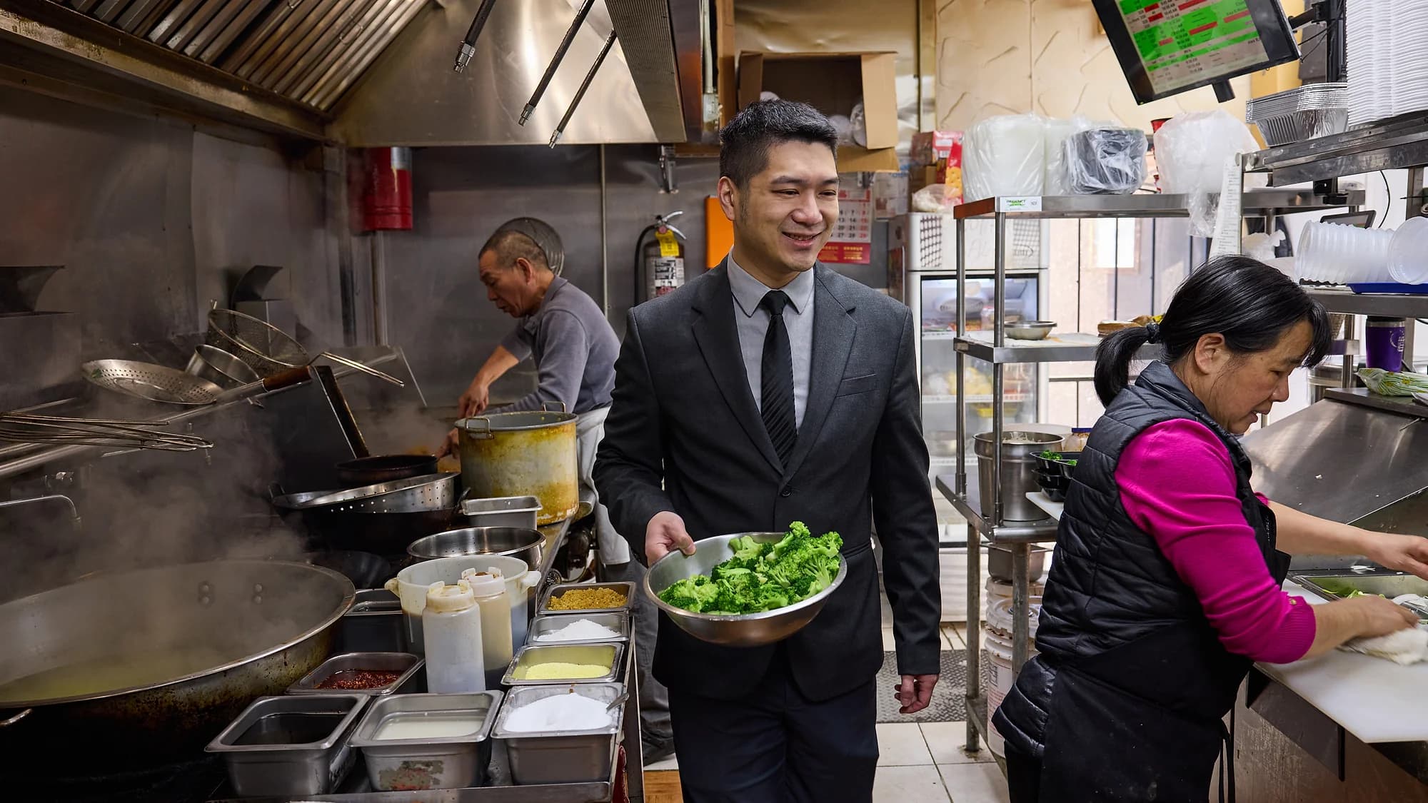Patrick Cheng carrying a bowl of fresh broccoli through the General Tso's kitchen while two cooks work at the wok station and the prep counter